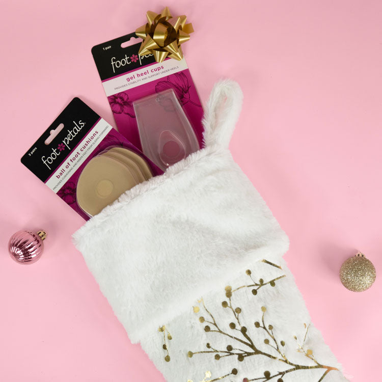 Foot Petals Ball of Foot khaki cushions and Foot Petals Gel Heel Cup cushions in white furry stocking on pink background next to holiday tree ornaments