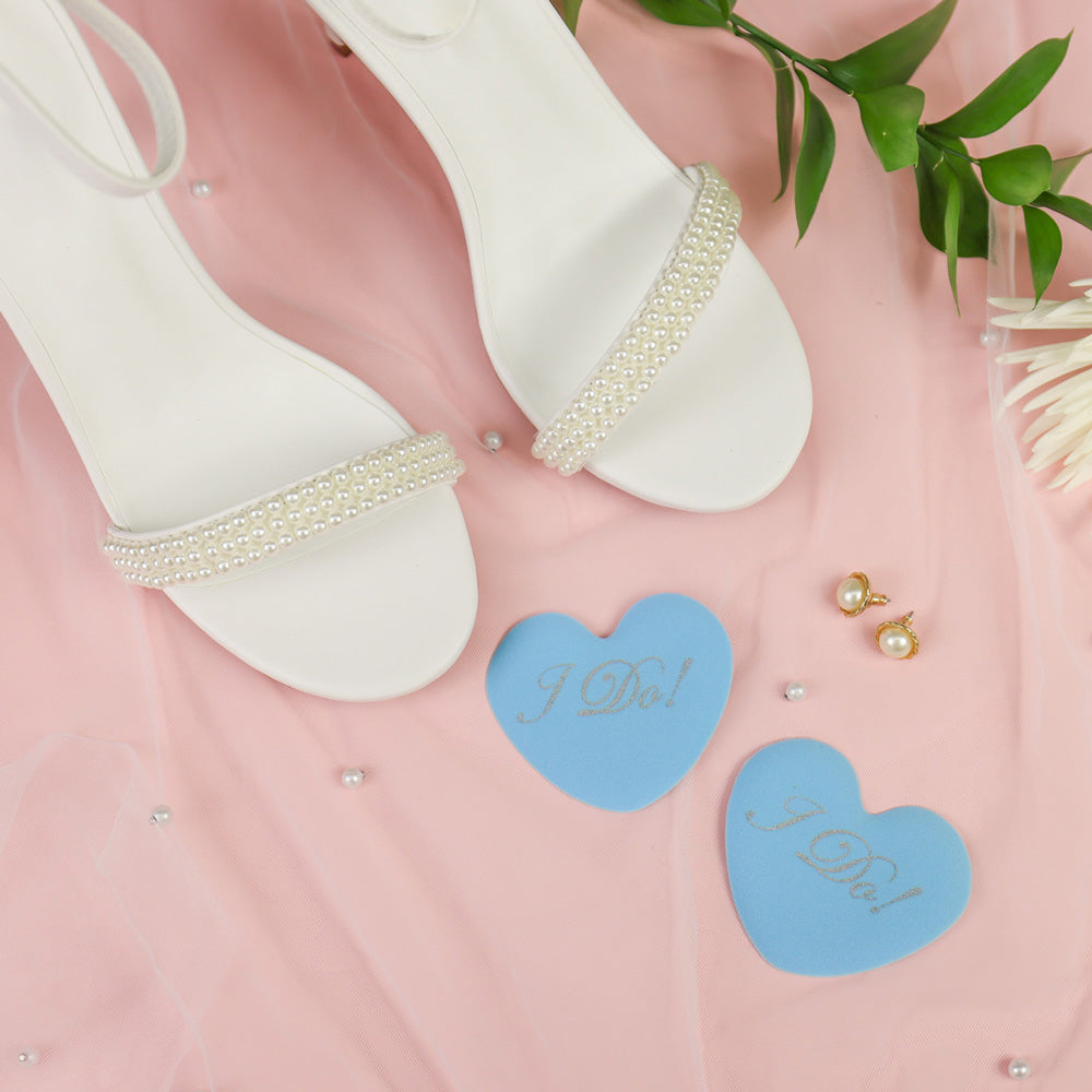 White sandals with pearl details on a pink surface with 'I Do!' heart cushions and earrings.