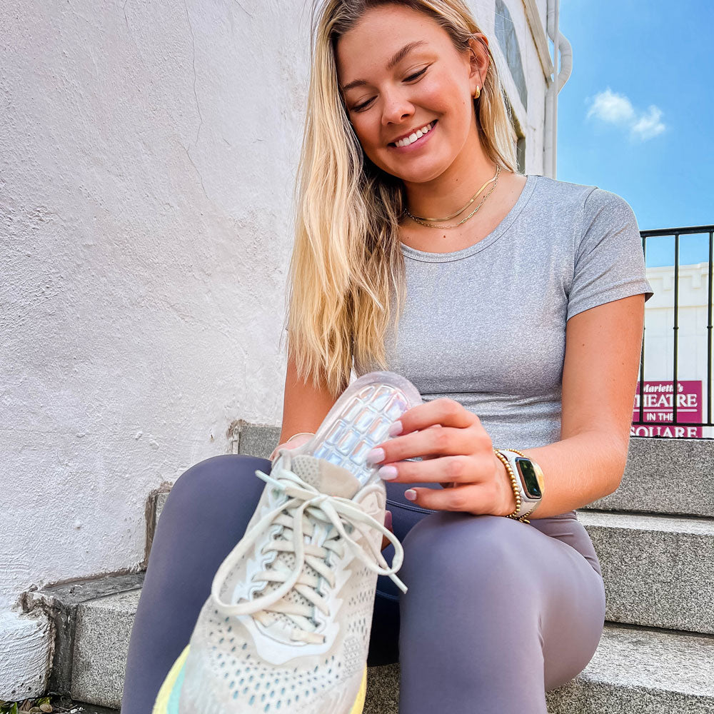 Woman sitting on concrete stairs while holding sneaker and placing Foot Petals Air Gel Heel Cup cushions inside.