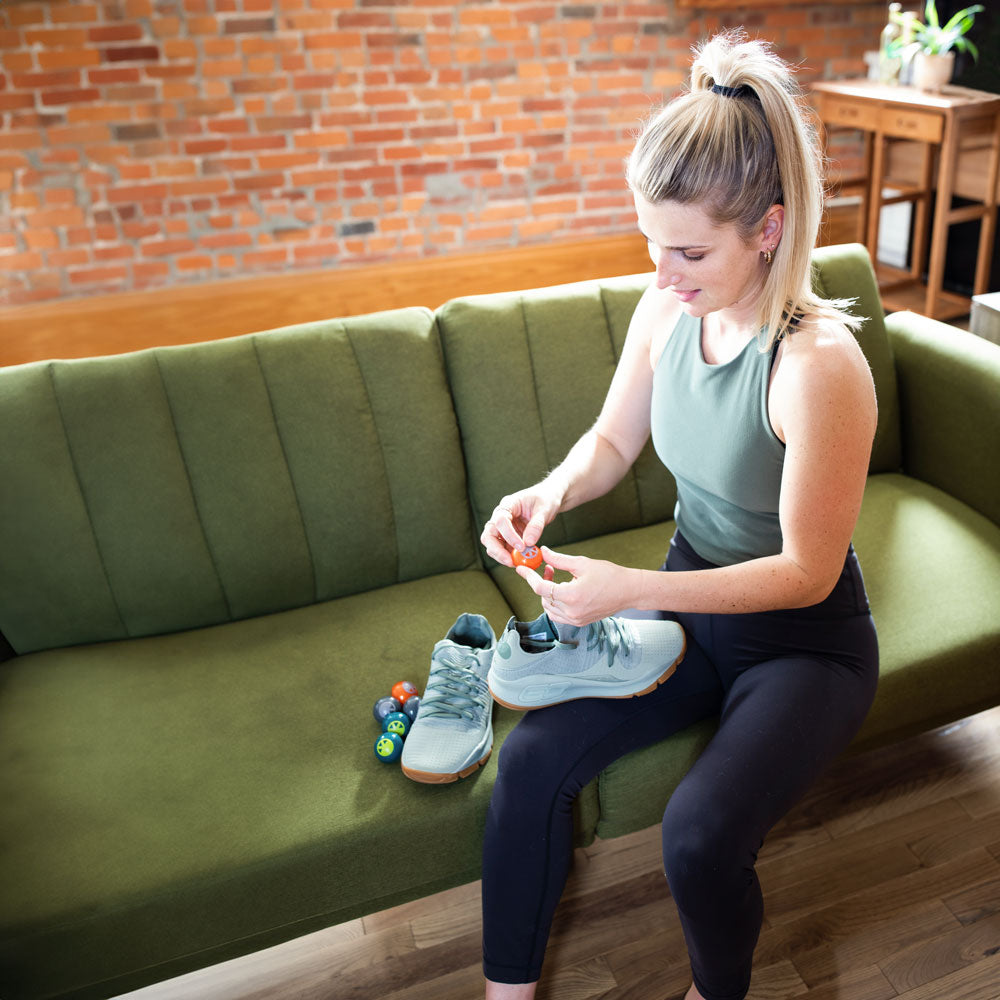 Woman sitting on green sofa in living room while placing Foot Petals Sneaker Deodorizers into light blue sneakers #color_neutral-3-pairs